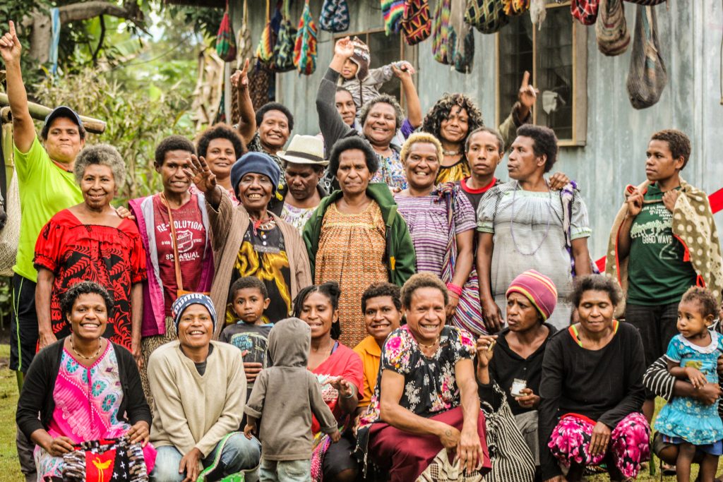 A group of 20-25 women weavers and a few children smile joyfully at the camera. Colourful bilum bags hang along the side of a building behind them.