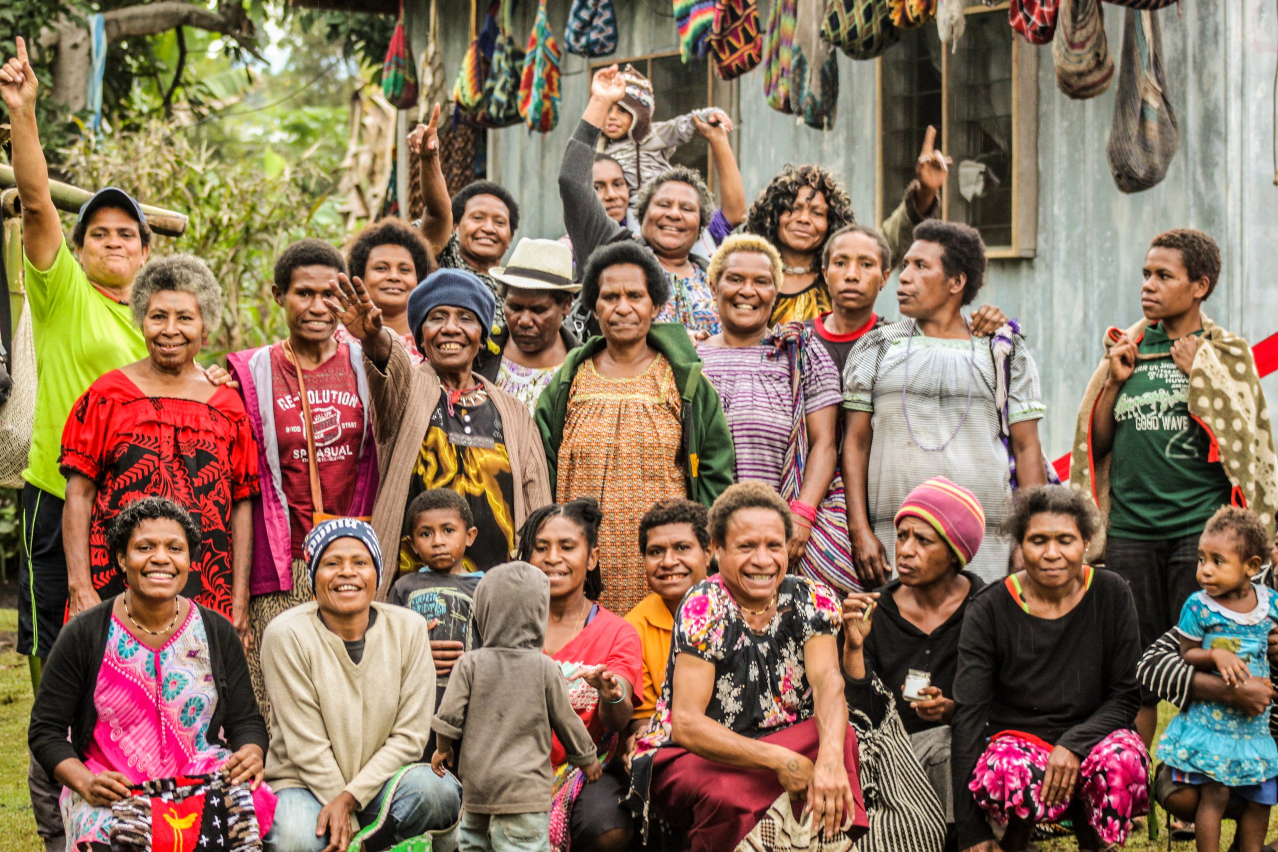 group of 20-25 women weavers and a few children smile joyfully at the camera. Colourful bilum bags hang along the side of a building behind them.